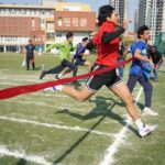 Students sprint to the finish line in a school race, with one breaking the ribbon as others follow closely.
